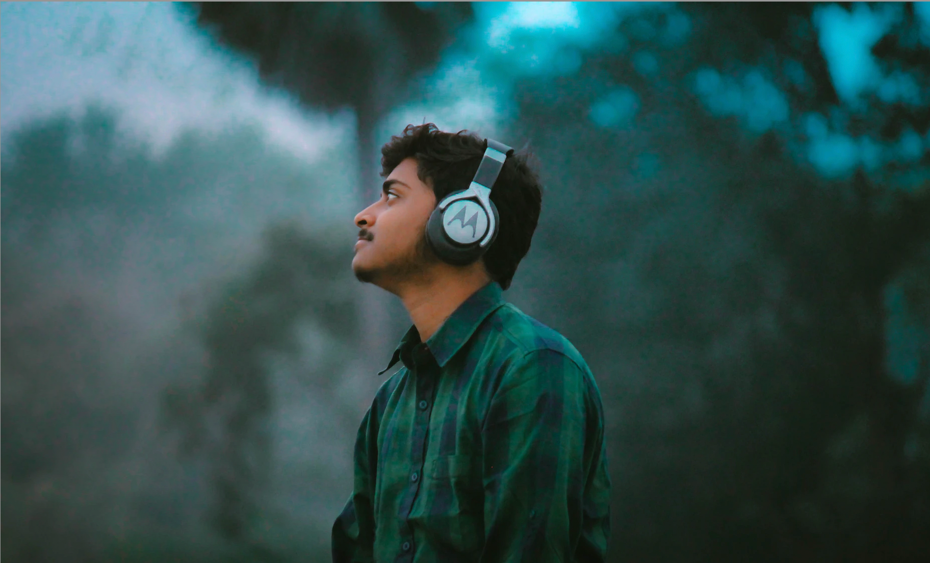 A man with headphones on stares thoughtfully into the distance. A forrest of trees is out of focus in the background.