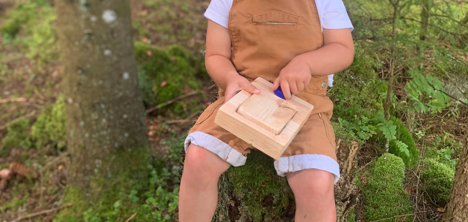 Close up of a young child playing with a wooden Voxblock prototype. They are sitting on a tree stump, and surrounded by greenery.