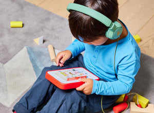 5 year old boy holding a voxblock player with red bumper, listening to an audiobook using green headphones