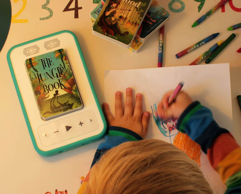 Child drawing a picture with a voxblock player with green bumper on the table. The audiobook he is listening to is the Jungle Book which has a green cover with plants. 