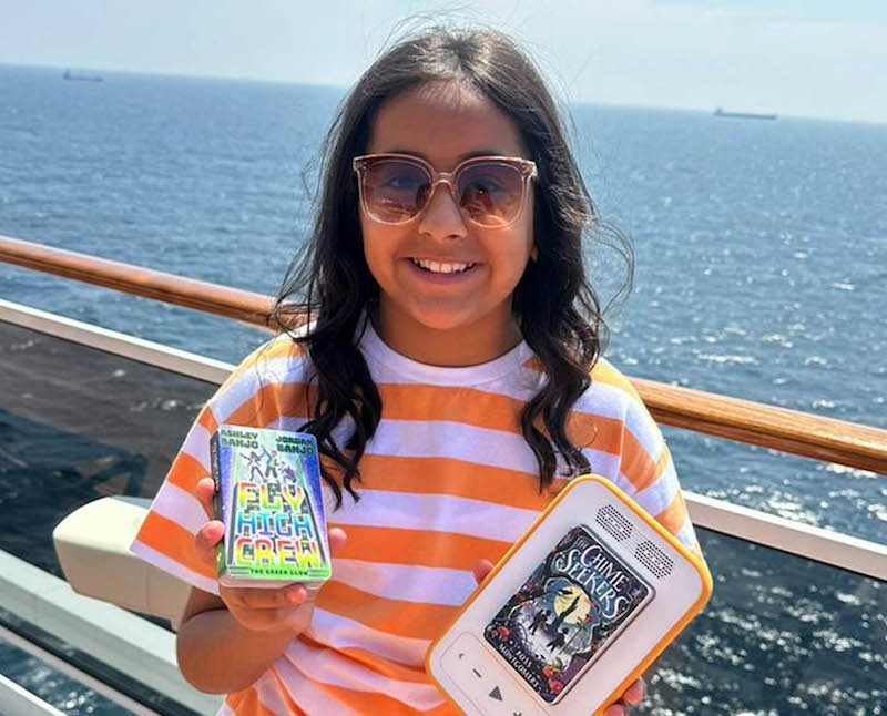 10 year old girl smiling on holiday holding a voxblock player in one hand and an audiobook in the other. 