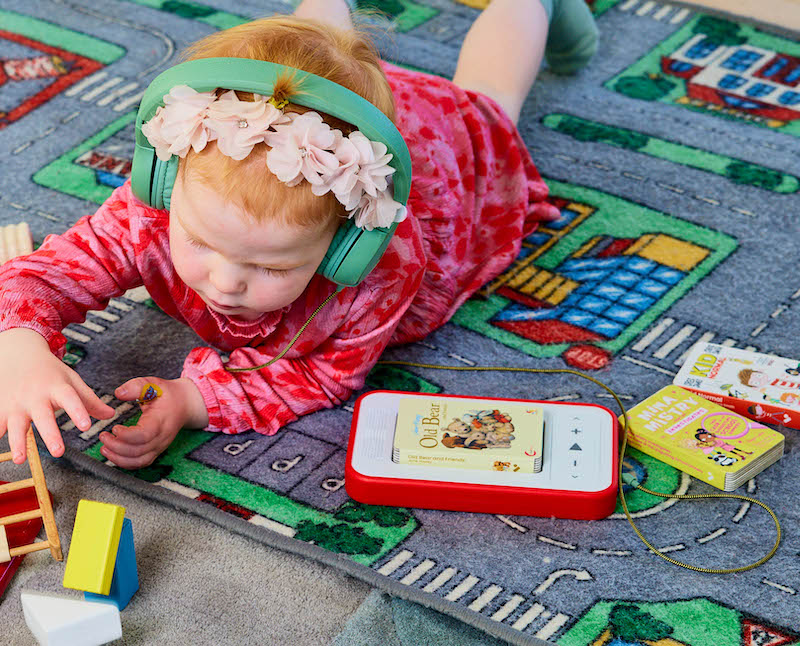 Young girl lying down playing with wooden toys whilst listening to a voxblock audiobook player using green headphones