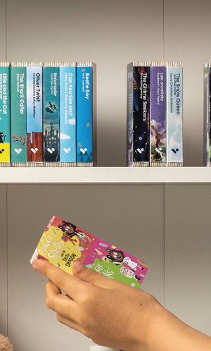 A hand taking the Maggie Sparks and the Truth Dragon & Maggie Sparks and the School of Slime audiobook from a shelf of Voxblock audiobooks