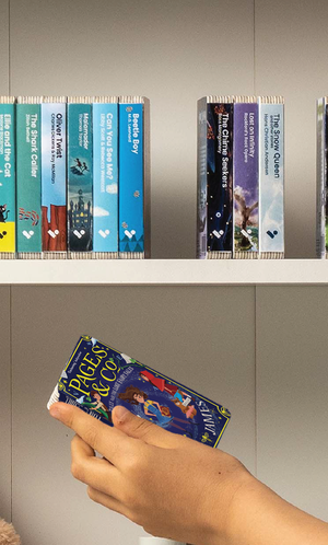 A hand taking the Pages & Co.: Tilly and the Lost Fairy Tales audiobook from a shelf of Voxblock audiobooks