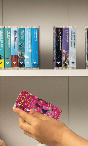 A hand taking the Pages & Co.: Tilly and the Map of Stories audiobook from a shelf of Voxblock audiobooks