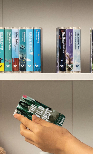 A hand taking The Wild Robot audiobook from a shelf of Voxblock audiobooks